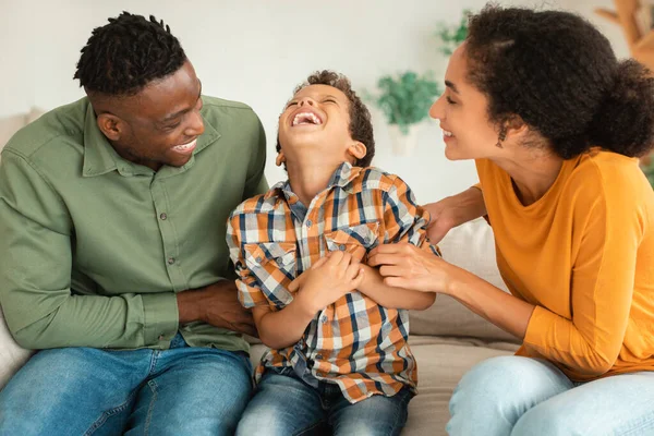 Parents And Kid Bonding Fun. Cheerful Multicultural Family Playing And Laughing, Dad And Mom Tickling Little Son Sitting On Couch At Home Interior, Bonding And Enjoying Time Together On Weekend