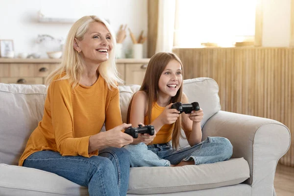 Emotional happy caucasian family senior lady grandmother and girl teenager granddaughter sitting on couch, holding joysticks consoles in their hands, playing video games at home, copy space