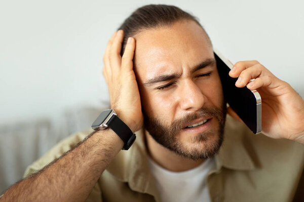 Closeup of millennial man in pain touching his head and calling doctor or ambulance on phone, suffering from migraine, copy space. Young guy have phone conversation, hear bad news