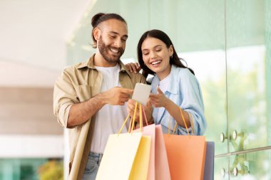 Bir sürü paketi olan gülümseyen beyaz bir çift dışarıda gezmekten, alışveriş yapmaktan, akıllı telefon uygulaması ve online bankacılık kullanmaktan zevk alıyor. Tavsiye satışı, nakit para, yaşam tarzı, reklam ve teklif