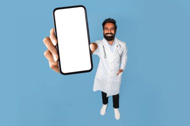 Tıbbi uygulama. Üniformalı Hintli erkek doktor, elinde boş bir ekranla akıllı telefon, elinde telefon olan doktor, mavi arka planda duruyor,
