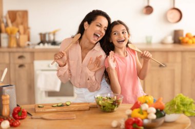 Aile Aşçılığı Eğlencesi. Neşeli anne ve kızı kaşıklarla şarkı söylerken modern mutfağın yanında organik sebzelerle birlikte taze salata hazırlıyor.