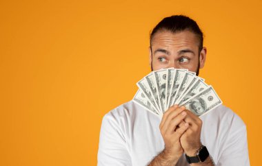 Komik gülümseyen yetişkin Avrupalı adam elindeki bir sürü doların fotokopi alanına bakıyor, turuncu stüdyo arka planında izole edilmiş. Kâr, reklam ve teklif, tasarruf finansmanı, kazanç ve zenginlik