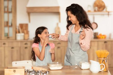 Yemek pişirme eğlencesi. Gülümseyen anne ve kızı, modern mutfakta birlikte yemek pişirirken, anne un içinde ellerini tutarken ve hafta sonları pasta yaparken çocukların burnuna dokunurken.