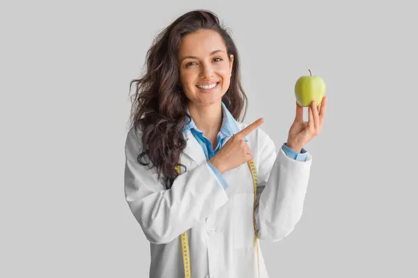 身穿制服的年轻女医生兴高采烈地把手指放在被浅灰背景隔开的苹果上 对着相机笑着 营养学家的建议 — 图库照片