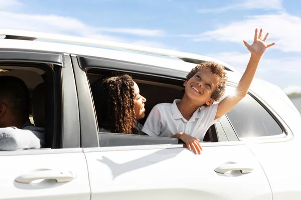 Happy african american mother and child looking from white car window, waving and smiling, enjoying view. Cheerful black family enjoying road trip by brand new auto, going vacation, weekend journey