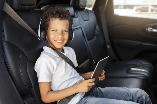 Cheerful cute african american school aged boy sitting at car back seat, using wireless headphones and digital tablet, playing video games while going home from school, smiling at camera, copy space