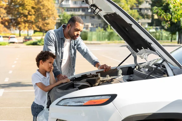アフリカ系アメリカ人の父親と子供が駐車場に立って 壊れた自動車のエンジンを修理しています 若い黒人の父親は 家族の車のいくつかの部分を修理するために彼の息子を教えています — ストック写真