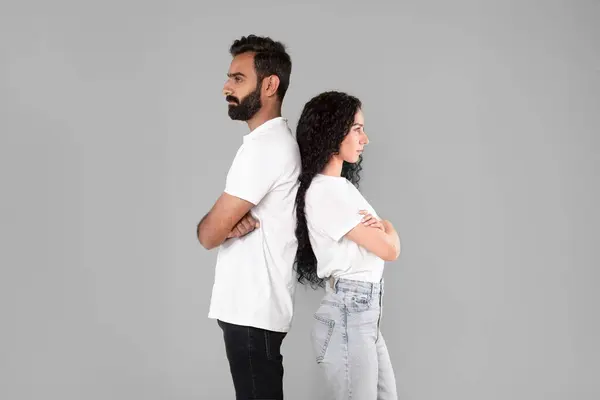 Relationship crisis and quarrels. Offended middle eastern couple ignoring each other after conflict, standing back to back crossing hands on gray background. Spouses suffering from marriage issue