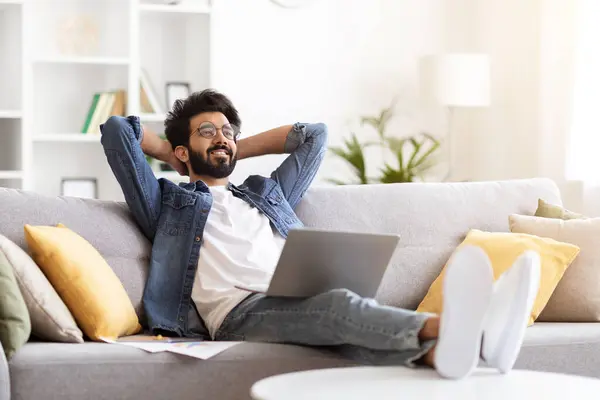 Portrait Of Relaxed Young Indian Man With Laptop Sitting On Couch At Home Interior, Smiling Eastern Freelancer Guy Leaning Back On Couch With Hands Behind Head, Having Break In Online Work