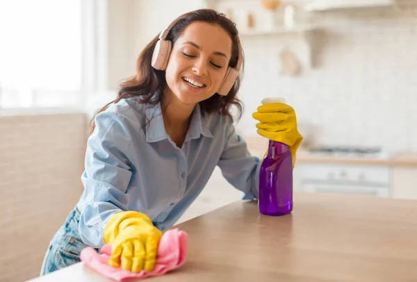 Excited woman cleaning table in kitchen from dirt and listening music in headphones, lady using rag and spray detergent for polishing surface, enjoying doing domestic chores