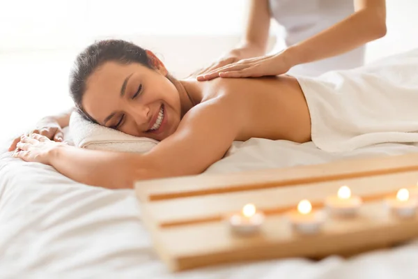 Young Woman Relaxing In Spa Salon During Massage Session Indoor, Unrecognizable Masseur Rubbing Her Back While Lady Lying On Table Near Burning Candles And Enjoying Beauty Treatment At Spa Center
