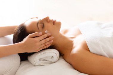 Kapalı gözlerle uzanan, havlunun üzerinde dinlenen ve güzellik prosedürünün tadını çıkaran bir kadın olarak kaplıcada kadınların kafalarına ve tapınaklarına masaj yapan ellerin yakın çekimi. Seçici odak