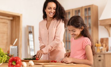 Aile yemekleri. Mutlu çocuk kızı ve annesi mutfakta akşam yemeği için salata hazırlarken taze organik sebzeleri kesen bir kıza odaklanıyorlar. Sığ Derinlik