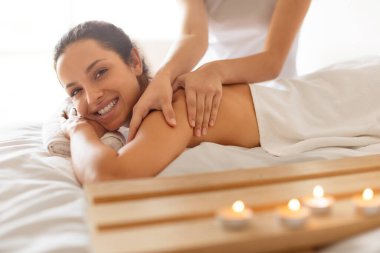 Smiling young lady having full body massage lying on table at spa salon, looking at camera, enjoying beauty treatment while therapist rubbing her shoulders and back indoors. Bodycare and wellness clipart