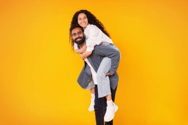 Pozitif Ortadoğulu genç eşler poz verirken eğleniyor, koca eşini sırtında taşıyor, sarı arka planda ona biniyor ve ona biniyor, stüdyoda çekim yapıyor. Birlikte Mutlu Kavram