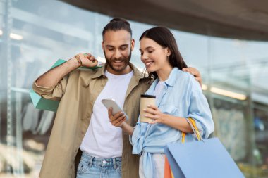 Bir sürü çantası olan neşeli beyaz kadın ve koca akıllı telefon uygulaması kullanarak yürüyüş, açık hava, online alışveriş yapmaktan zevk alıyor. Satış, nakit para, alışveriş hayatı, kazan, reklam ve teklif.