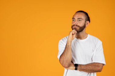 Beyaz tişörtlü sakallı düşünceli, düşünceli, akıllı, yetişkin Avrupalı adam, turuncu stüdyo arka planında izole edilmiş boş alana bakıyor. Fikir yarat, yaşam tarzı seçimi, insani duygular yarat ve öner