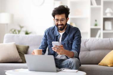 Sırıtkan Hintli genç adam evde koltukta otururken akıllı telefon kullanıyor ve kahve içiyor, cep telefonuyla mesaj atan ve dizüstü bilgisayarla çalışan mutlu Doğulu serbest yazar,