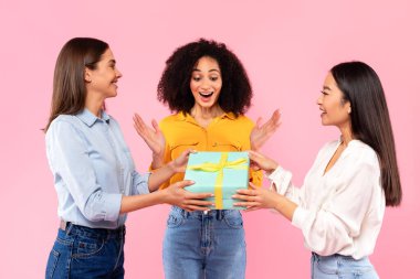 Tatilde ya da doğum gününde arkadaşlarından hediye alan heyecanlı siyahi kadın, çeşitli dişiler sürpriz bir bayana hediye veriyor, pembe stüdyo arka planının başında duruyor.