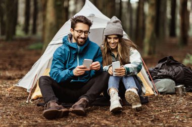 Ceketli, çadırın yanında oturan, akıllı telefondan sohbet eden, maceranın tadını çıkaran, hafta sonları boş vakit geçiren soğuk ormanda seyahat eden beyaz bir aile. Birlikte tatil, yaşam tarzı.