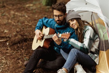 Çadırın yanında oturan, gitar çalan, müzikle eğlenen, sonbaharda soğuk ormanda hafta sonu geçiren beyaz bir kadın ve adam. İlişki, birlikte tatil, yaşam tarzı, macera.