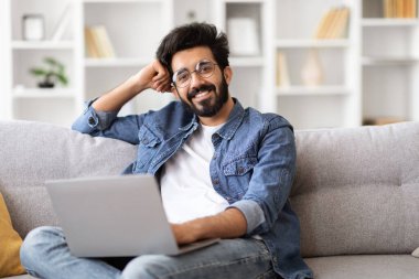 Gülümseyen genç Hintli erkek portresi dizüstü bilgisayarla evde koltukta dinleniyor, mutlu, yakışıklı, gözlüklü, koltukta oturan ve kameraya bakan ev içi rahatlığının keyfini çıkarıyor.