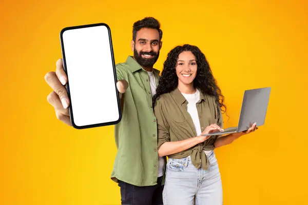 Online Offer. Cheerful Arabic Couple Posing With Big Blank Smartphone And Laptop, Man Showing Screen Recommending Mobile Application Over Yellow Studio Background. Check This App. Mockup