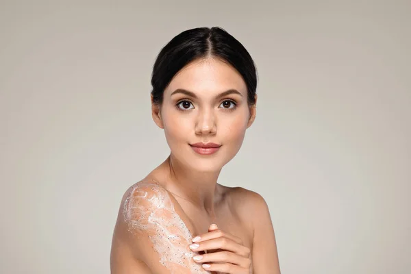 Happy calm millennial european woman with perfect skin applies cream, scrub to shoulder enjoy routine procedures at home, isolated on gray studio background. Body, beauty, skin care, spa treatment