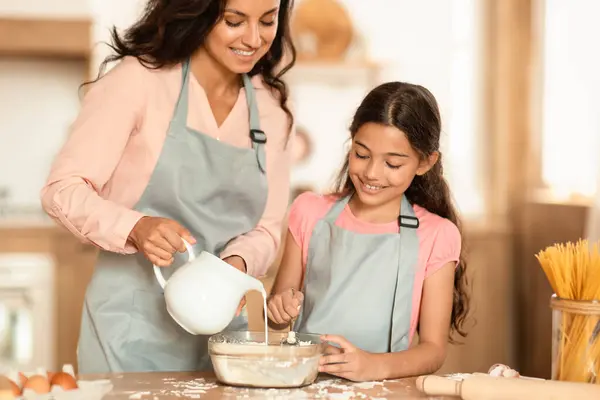 Smiling Mom And Preteen Daughter Baking Making Dough For Cookies, Adding Milk In Bowl Standing In Home Kitchen, Wearing Aprons. Family Pastry Recipes Concept. Cropped Shot