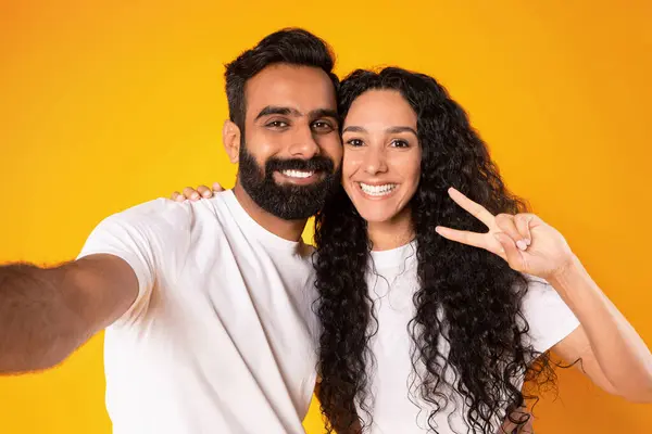 Peace And Love. Portrait of smiling arab couple posing for selfie, lady gesturing victory sign to camera embracing husband over yellow background. Studio portrait shot of positive spouses