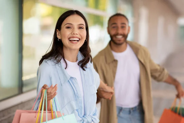 Happy young caucasian woman leads her husband by hand with many packages, enjoy huge sale, shopping and walk, outdoor. Shopaholic emotions, buy, recommendation sale, ad and offer
