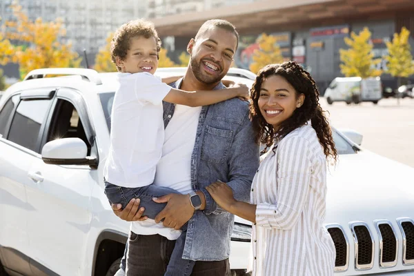Outdoor portrait of cheerful black family posing by their car at parking spot. Happy african american father, mother and kid preteen son going to shopping mall at weekend, time together