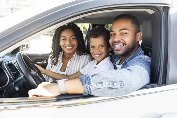 Happy black family with one child son riding car, spending time together at weekend, smiling at camera. Cute african american preteen boy sitting on dad lap, learning how to drive auto
