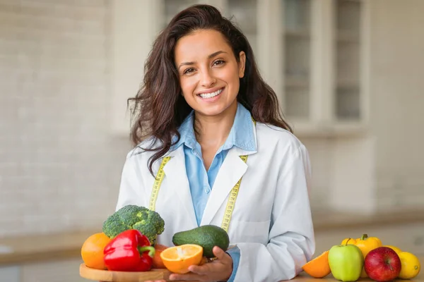 Portrait of smiling female nutritionist with plate of fresh fruits, working at weight loss clinic, posing and smiling at camera. Healthy nutrition consultant recommending healthy eating