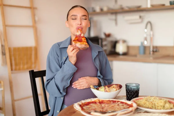 Pregnancy and cheat meal. Young expectant woman having desire for junk food, biting slice of pizza, sitting at table full of dishes in kitchen interior