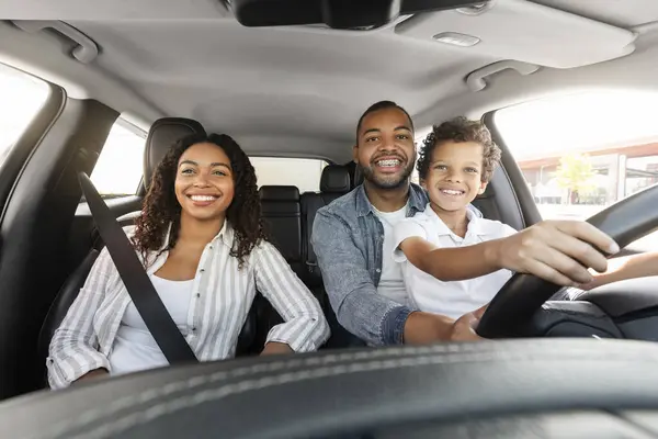 Happy millennial black parents father and mother enjoying brand new car with their child son. Excited preteen african american boy sitting on dad lap, driving automobile together and smiling