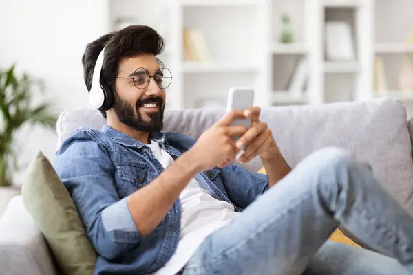 Home Leisure. Young Indian Guy In Wireless Headphones Relaxing With Smartphone On Couch, Listening Music And Messaging With Friends, Enjoying Domestic Pastime, Resting On Comfortable Sofa, Free Space