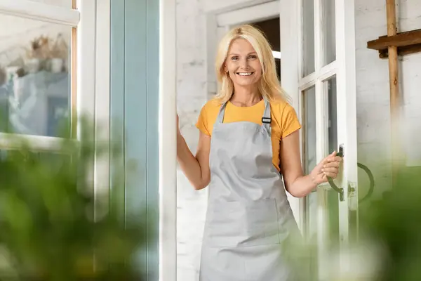 Happy Older Lady In Apron Opening Door And Welcoming Inside Her Country House, Smiling Mature Farmer Woman Standing At Terrace, Beautiful Older Female Looking And Smiling At Camera, Copy Space