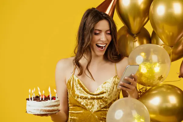 Excited lady in dress and birthday hat holding cake and reading congratulations on cellphone, standing near air balloons on yellow studio background