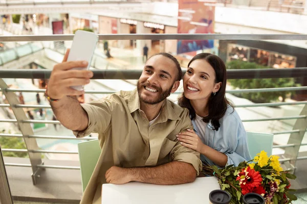 Happy young caucasian couple at table in mall cafe with flowers taking selfie on smartphone, enjoy date. Shopping, photos for social networks, relationships and lifestyle, sale, ad and offer