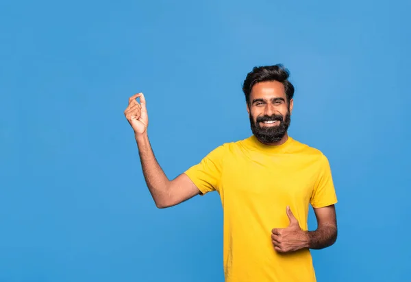 エキサイティングなインドの男は 手で目に見えないものを保持し 青いスタジオの背景の上に立って ジェスチャーを親指に見せます 広告のための空のモックアップ場所を示す肯定的なヒンドゥーの男性 — ストック写真
