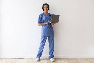 Sağlık ve Teletıp. Mavi önlüklü ve steteskoplu gülümseyen Hintli kadın doktor portresi. Dizüstü bilgisayar ve beyaz duvar arkasında duran doğu doktoru.