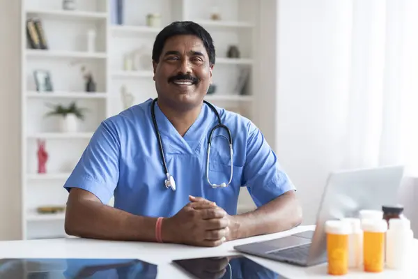 Smiling Indian Male Doctor In Blue Uniform Sitting At Desk With Laptop At Workplace, Professional Therapist Man Wearing Medical Scrubs And Stethoscope Posing In Office At Modern Clinic, Copy Space