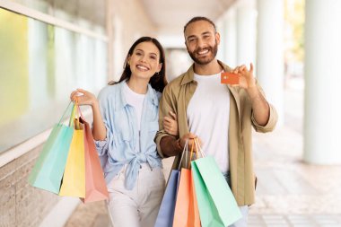 Bir sürü paketi olan gülümseyen beyaz bir çift dışarıda dolaşmayı, alışveriş yapmayı ve kredi kartını göstermeyi seviyor. Satılık finansman, reklam ve teklif, alışveriş kolik yaşam tarzı