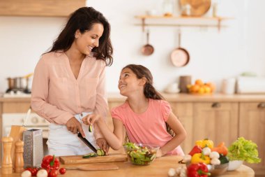 Aile yemeği hazırlığı. Neşeli anne ve küçük kızı modern mutfakta yemek pişirirken anne evde taze salata hazırlarken çocuk da salatalık dilimliyor. Beslenme Konsepti