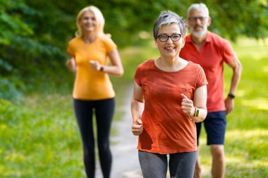 Bir grup aktif kıdemli insan parkta birlikte koşuyor, gülümseyen olgun erkek ve kadınlar dışarıda kardiyo eğitimi alıyorlar, doğada koşuyorlar, emeklilikte sağlıklı aktif yaşam tarzının tadını çıkarıyorlar.