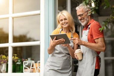 Modern Teknolojiler. Önlüklü, gülümseyen, dijital tablet kullanan ve terasta dikilirken kahve içen olgun çiftçiler Mutlu eşler Gadget 'ta internete göz gezdiriyorlar.