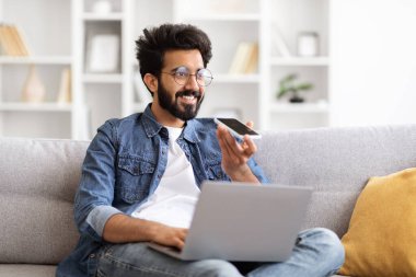 Genç Hintli adam evdeki sesli mesajı kaydetmek için akıllı telefon kullanıyor koltukta oturan, dizüstü bilgisayar kullanan ve cep telefonundaki sanal asistanı tarayan gülen doğulu adam,