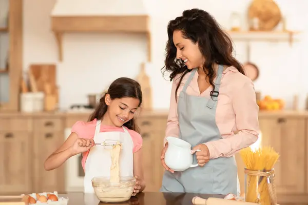 Cheerful Mom And Daughter Cooking And Baking Cake Together, Mother Looking At Happy Kid Girl Kneading Dough Mixing Ingredients In Bowl Standing In Kitchen At Home, Wearing Aprons